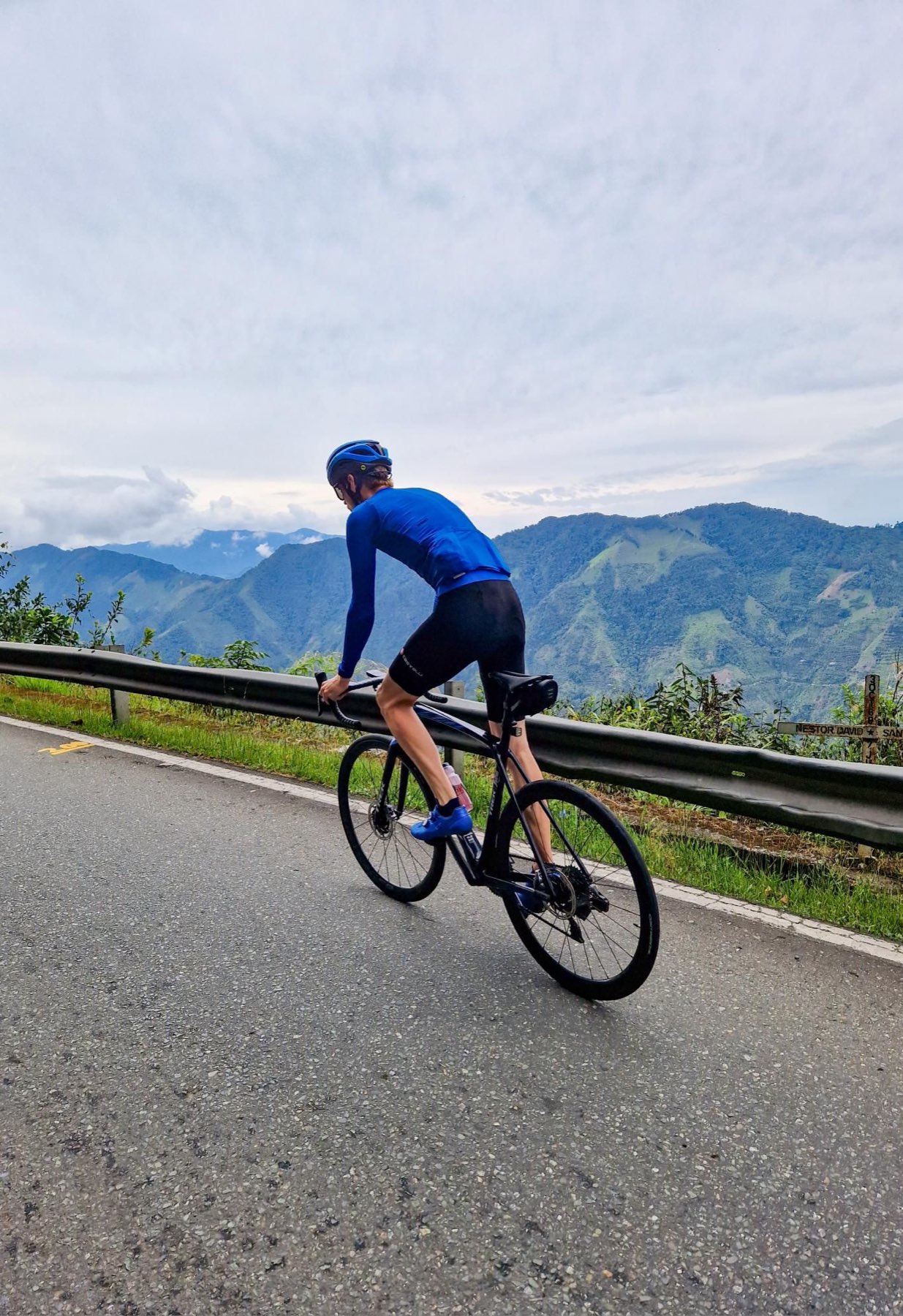 A solo cyclist climbing a guardrail-lined mountain road high above a hazy green valley
